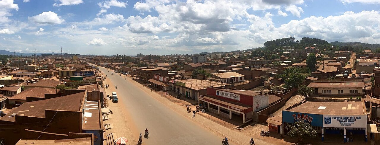 A main road through Butembo lined with shops, with motorbikes and pedestrians under a cloudy sky