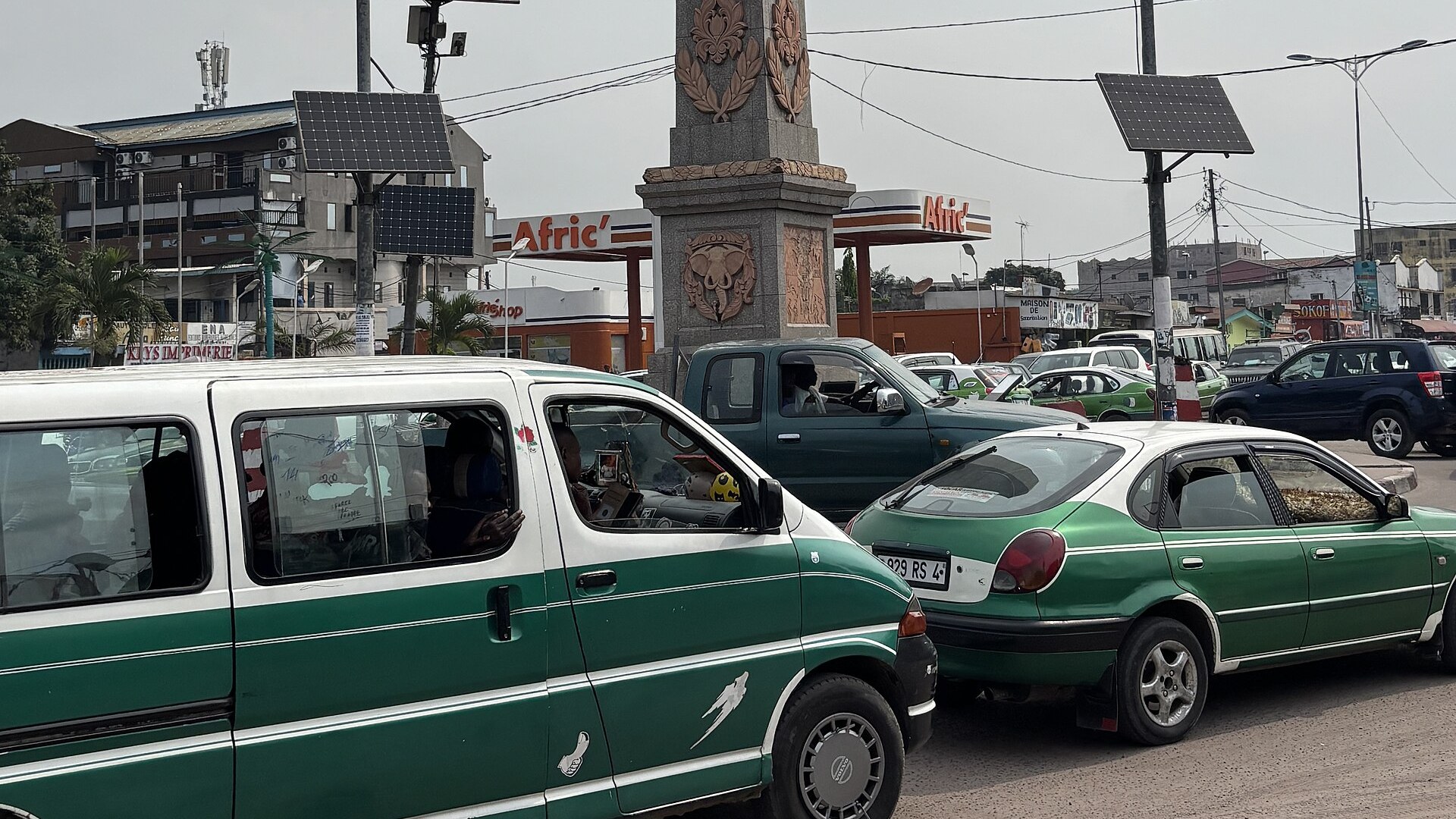 Green shared taxis queue at the Moungalie crossroads in Brazzaville, Republic of Congo, beside a stone obelisk