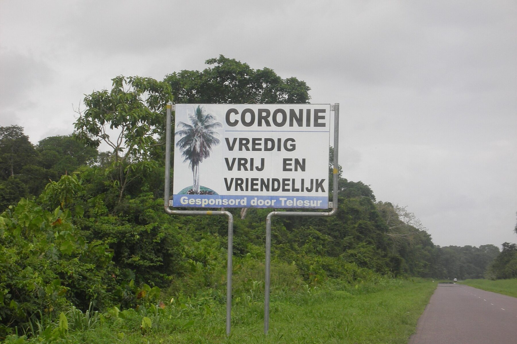 A white roadside sign reading Coronie, Vredig Vrij en Vriendelijk, with a palm tree logo, beside a two-lane road lined with dense green forest under a grey sky