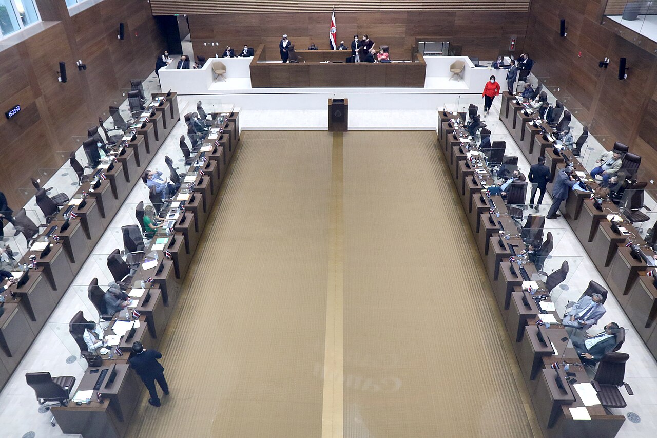 Deputies at their desks in the chamber of Costa Rica's Legislative Assembly, seen from the public gallery, with the national flag behind the presiding table