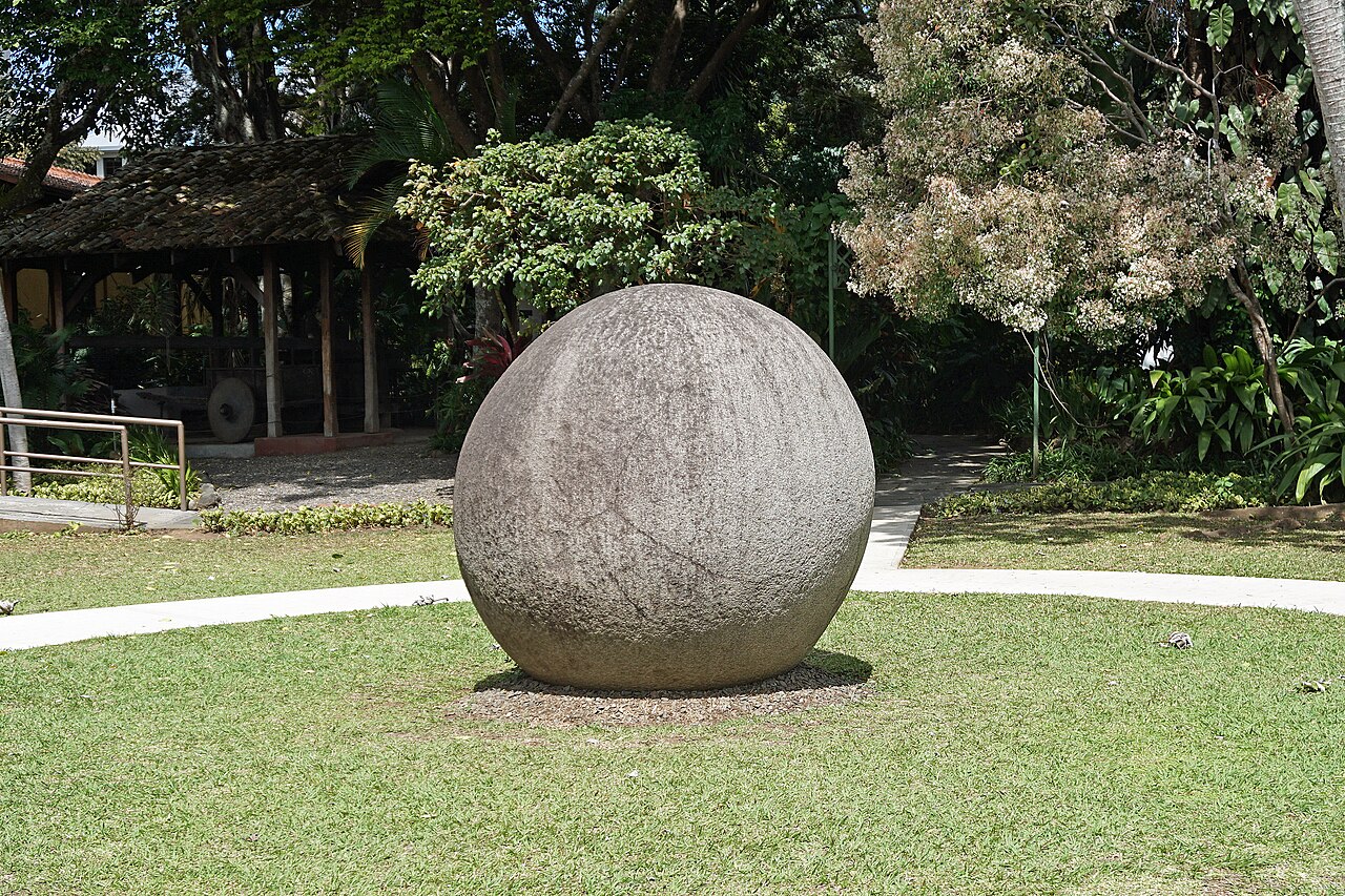 A large round grey stone sphere on a lawn in a garden, with trees and a thatched shelter behind it.