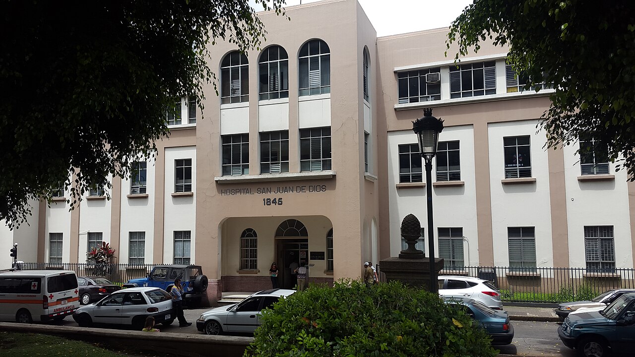 The beige façade of Hospital San Juan de Dios in San José, with 1845 above the arched entrance, an ambulance and parked cars in front
