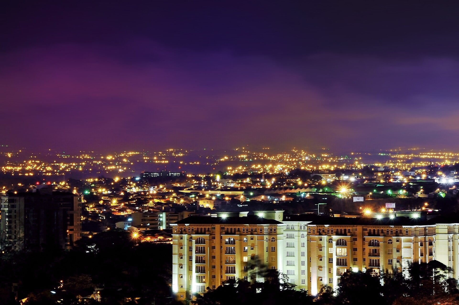 Night view of the Escazú area of San José province, Costa Rica, with city lights spread across the valley and a lit apartment block in the foreground under a purple sky