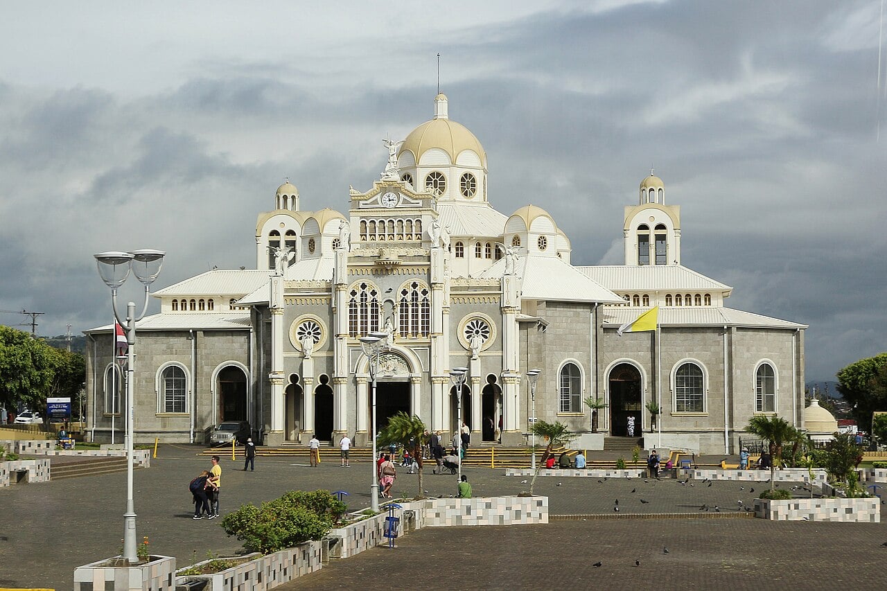 White and grey facade of the Basilica of Our Lady of the Angels in Cartago, with domes, a clock and a paved square in front