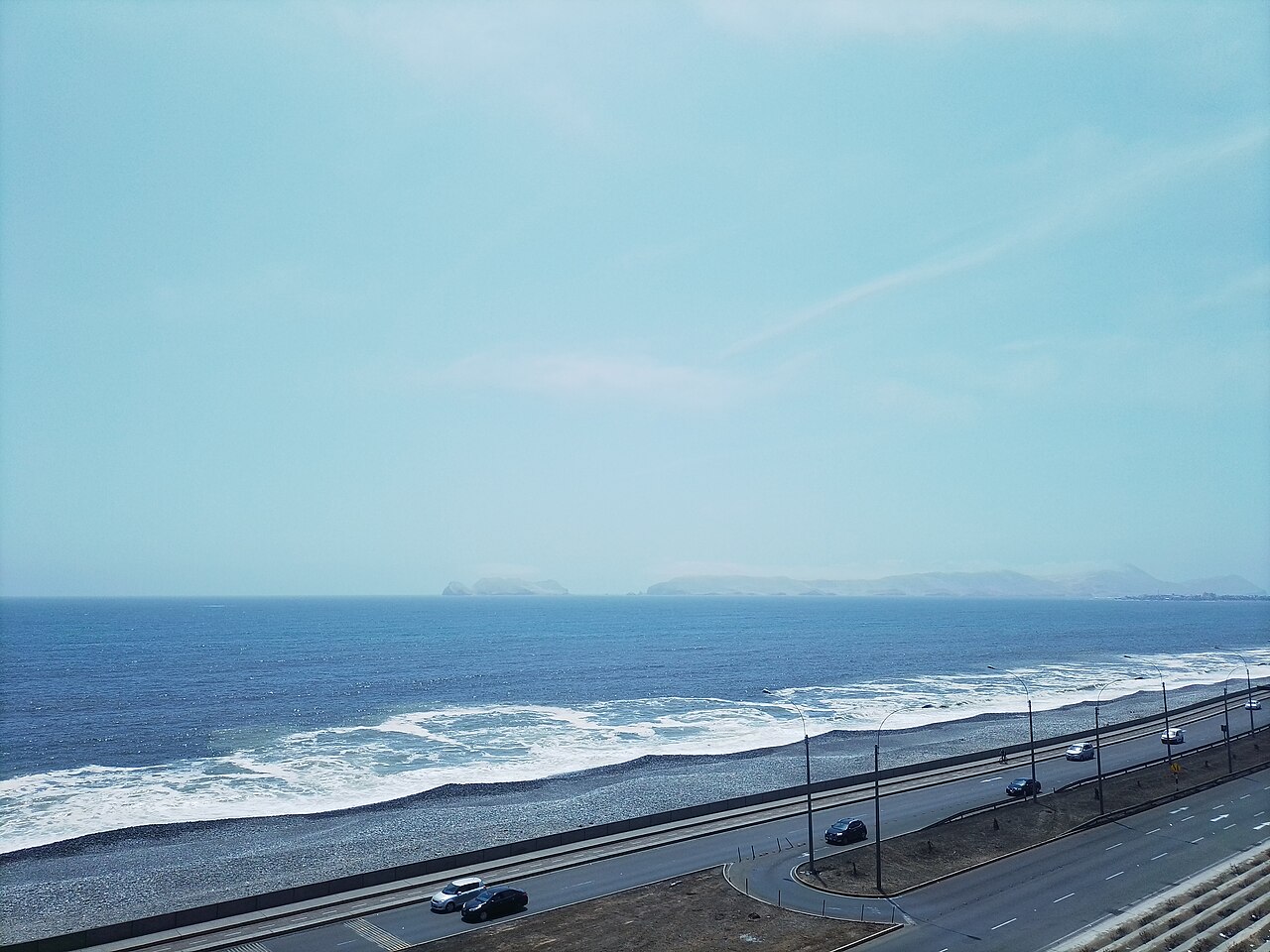The Costa Verde coast road and a pebble beach in San Miguel, Lima, with waves and a hazy Pacific horizon.