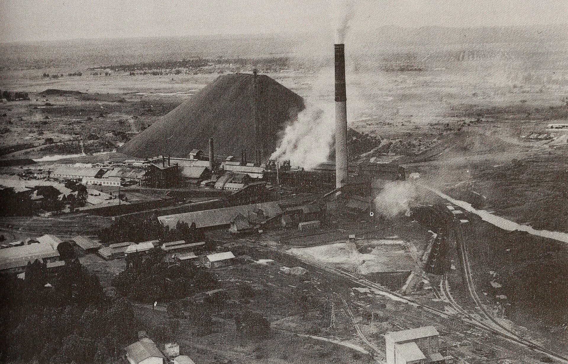 Black-and-white aerial view of the Gecamines copper smelter in Lubumbashi with a tall chimney and a large slag heap