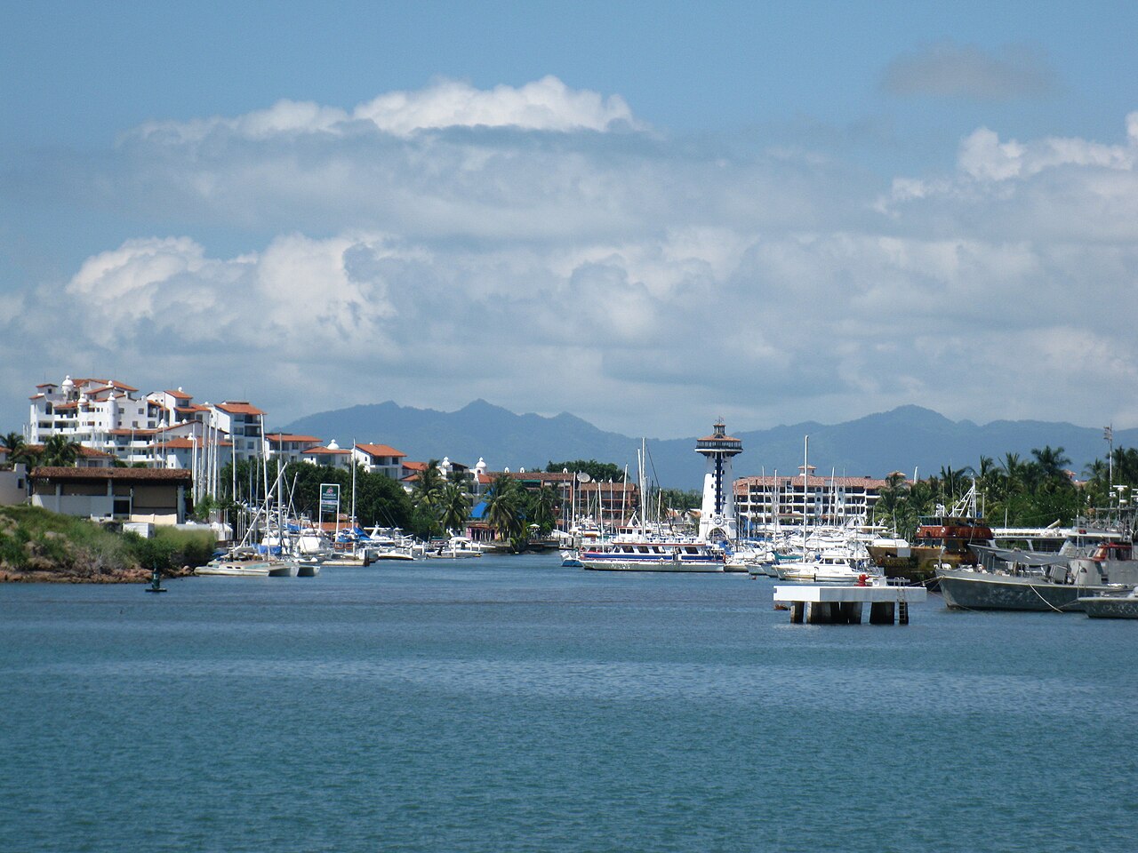 Yachts and tour boats moored in Marina Vallarta harbour, with a lighthouse tower, white buildings and mountains behind