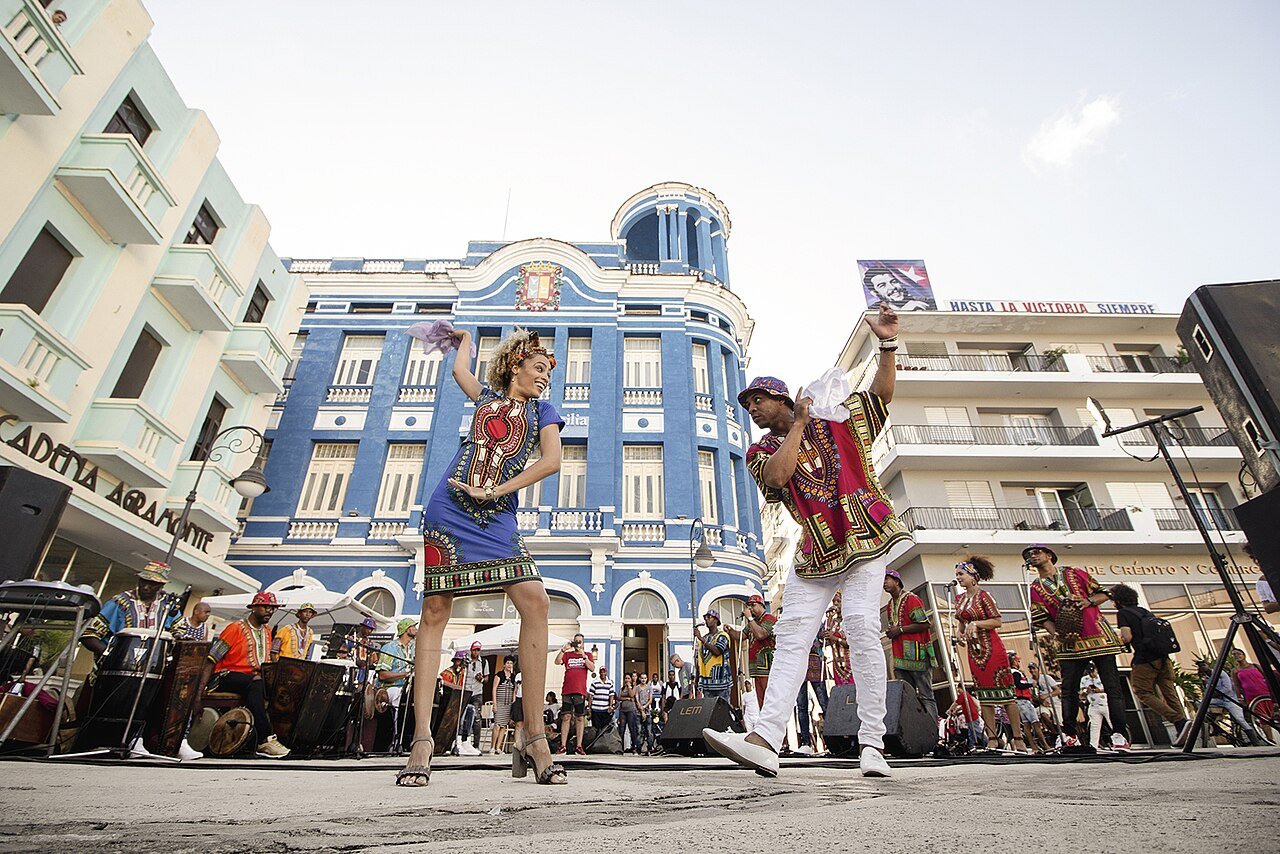 Two dancers in colourful African-print outfits perform Cuban rumba in front of a blue building while drummers play in a plaza in Camagüey, Cuba
