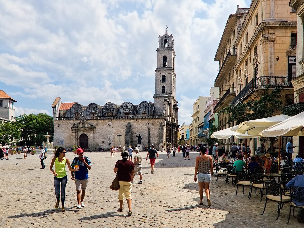People walking across a cobbled square in Old Havana past a stone church with a bell tower, beside a café terrace with white umbrellas