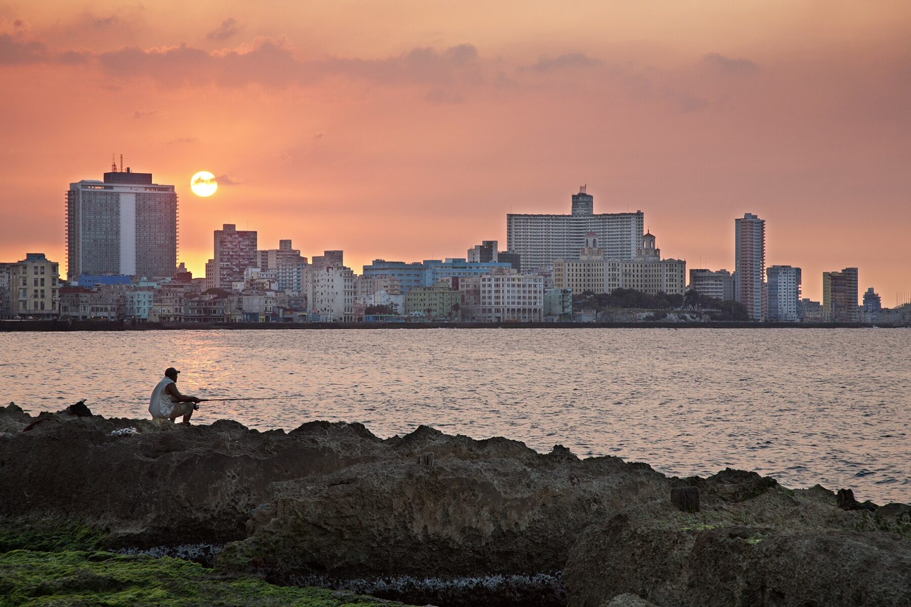 A man fishes from the rocks of the Malecón seafront in Havana, Cuba, at sunset with the Vedado skyline behind