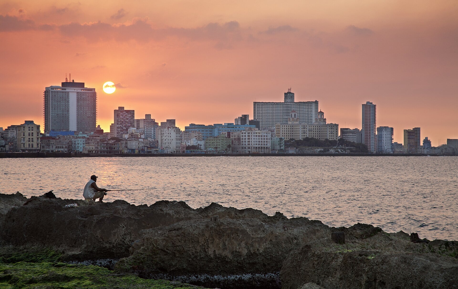 A man fishes from the rocks of the Malecón seafront in Havana, Cuba, at sunset with the Vedado skyline behind