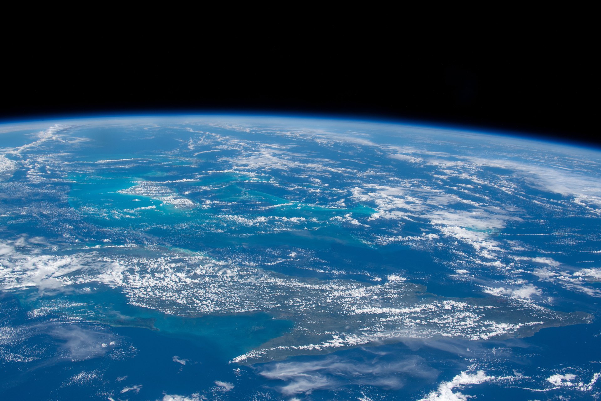 The island of Cuba seen from the International Space Station, with white clouds over green land and turquoise shallows under the curve of the Earth