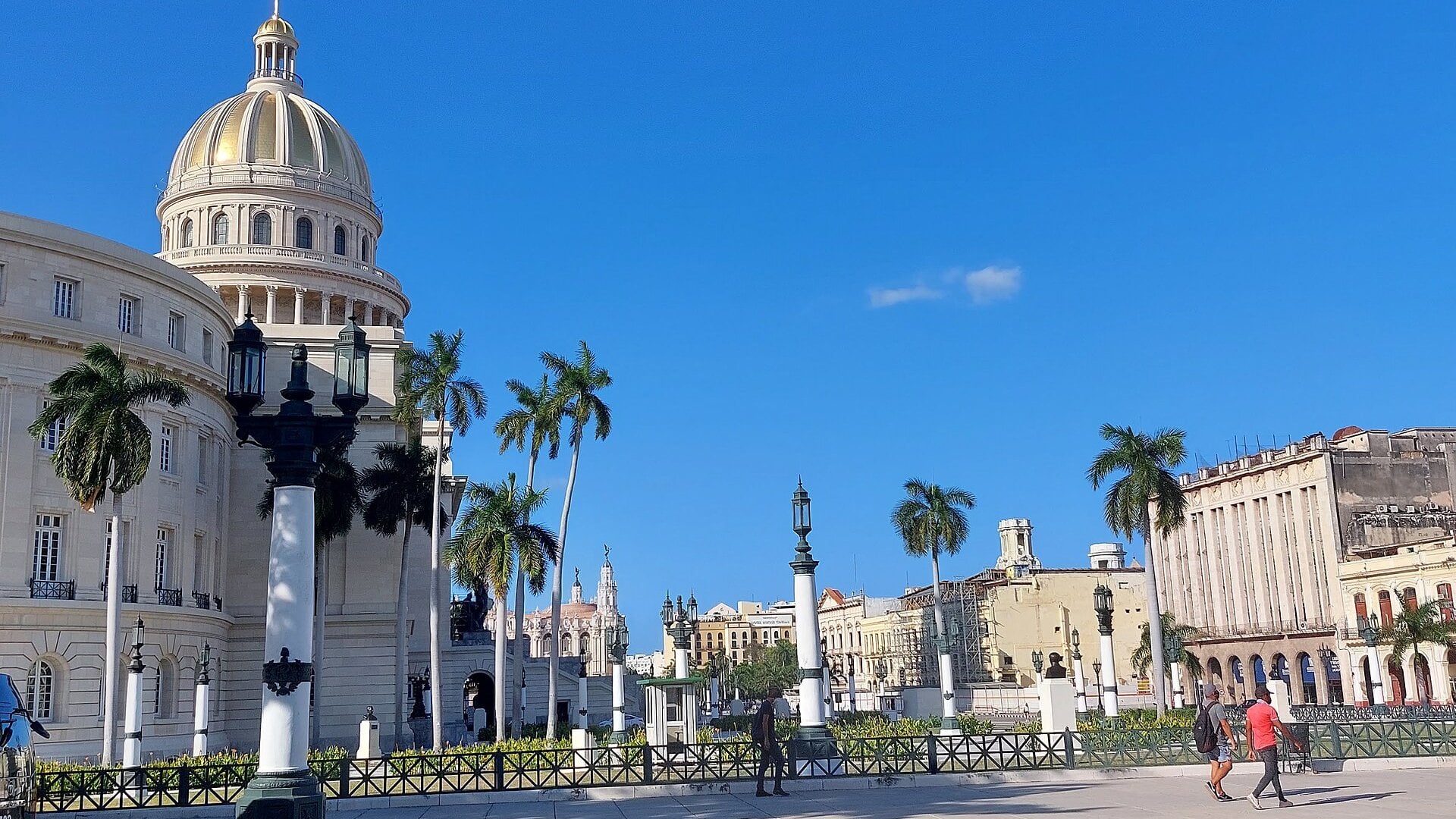 The domed Capitolio building in central Havana under a blue sky, with palm trees, street lamps and pedestrians in front