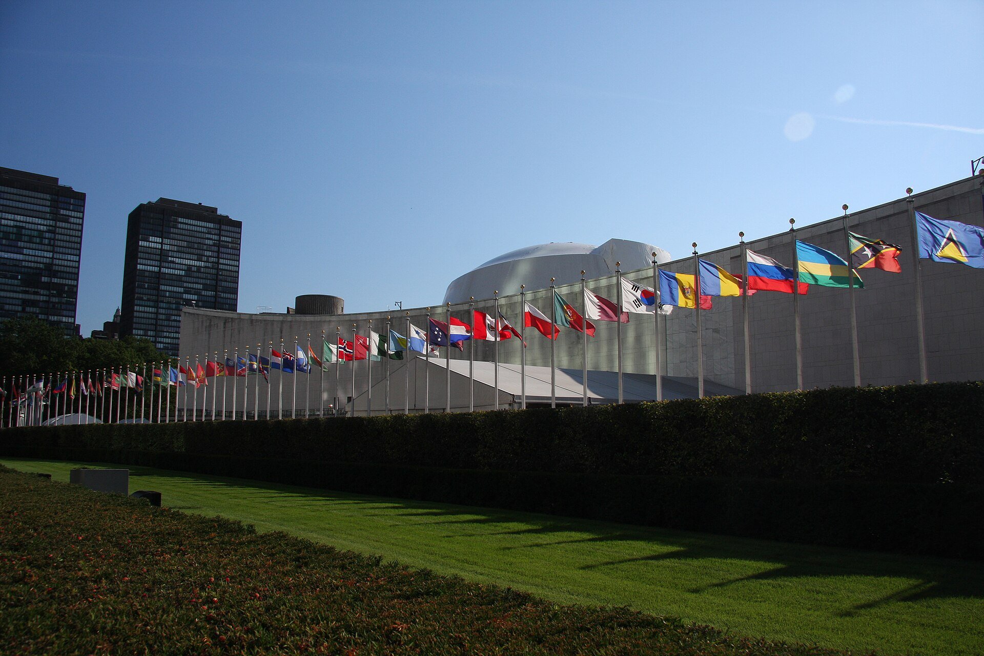 Member-state flags in a curved row in front of the United Nations General Assembly Building in New York under a clear blue sky