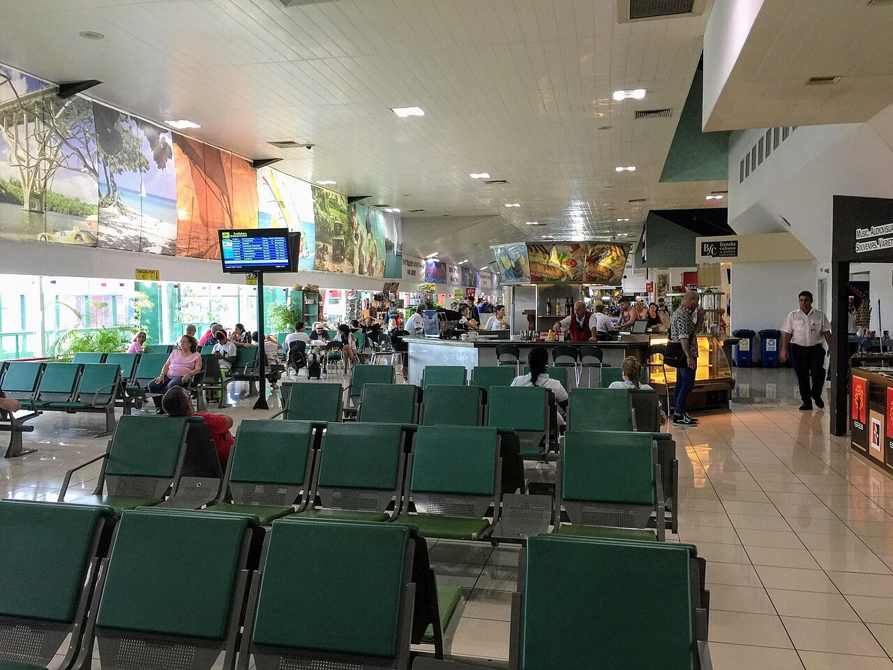 Rows of green seats in the departure lounge of Juan Gualberto Gómez International Airport in Varadero, Cuba, with a bar and a departures screen