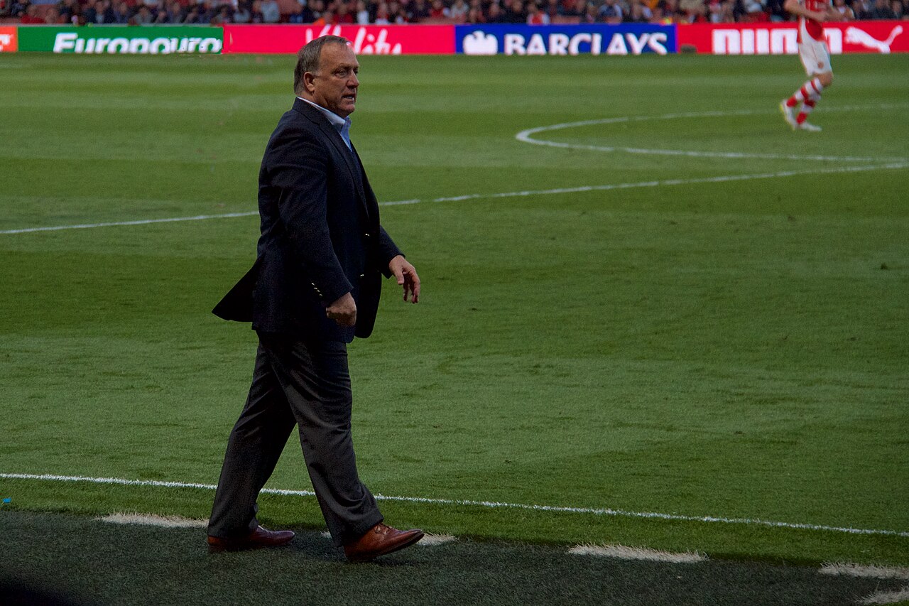 Dick Advocaat, grey-haired and wearing a dark blazer, grey trousers and brown shoes, walks along the touchline of a football pitch