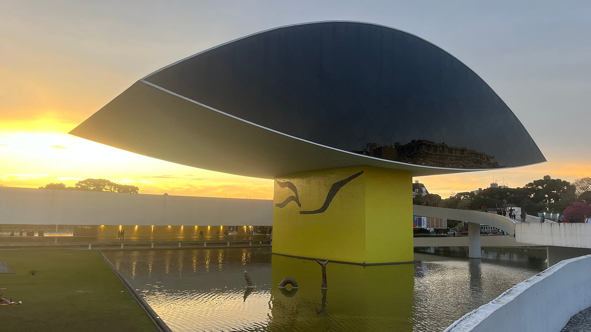 The Museu Oscar Niemeyer in Curitiba, Brazil, with its dark eye-shaped roof above a yellow pillar and reflecting pool at sunset
