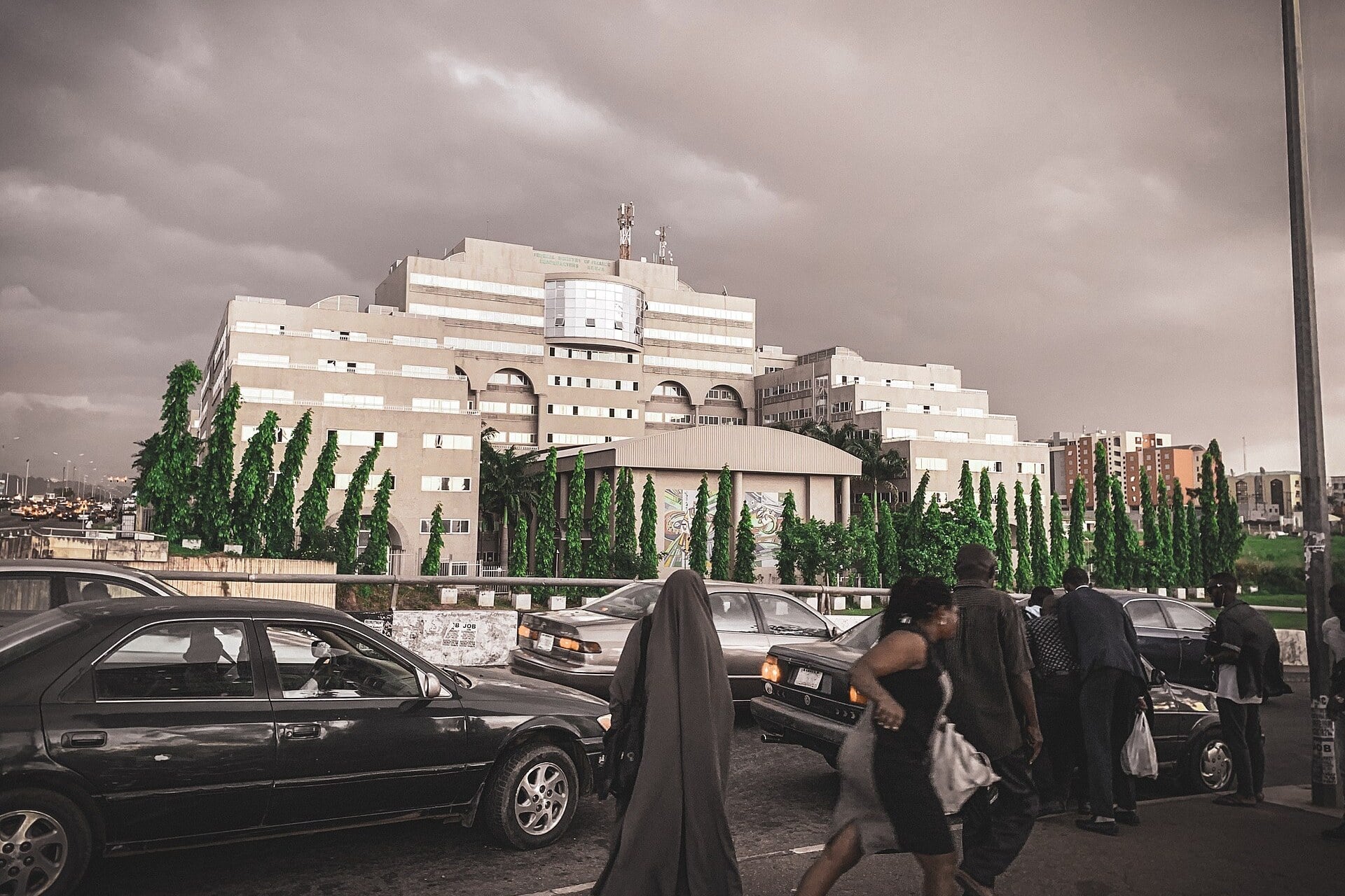 Pedestrians and cars in front of the Federal Ministry of Finance headquarters in Abuja under a cloudy sky