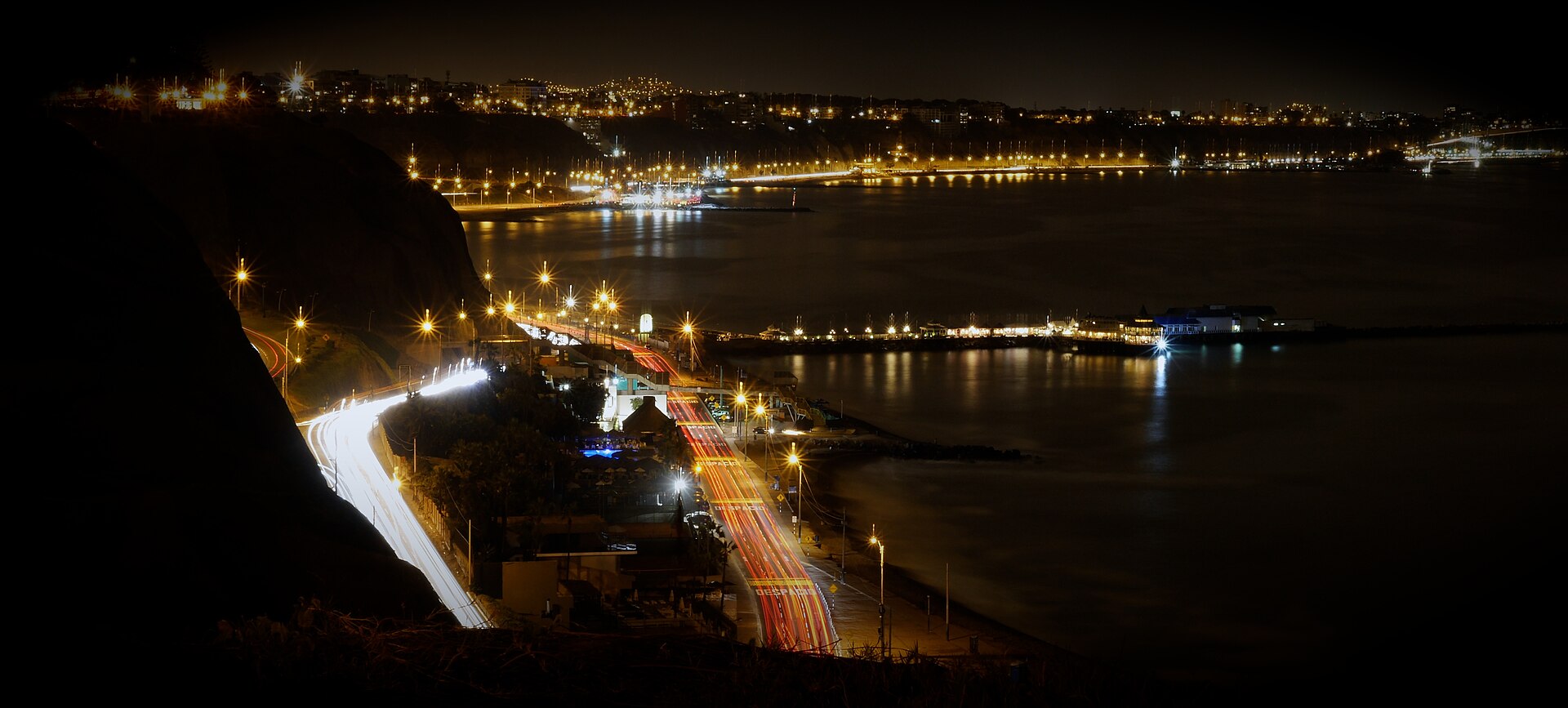 The Costa Verde coast road in Lima at night, with light trails from traffic beside a dark bay and the city lights beyond