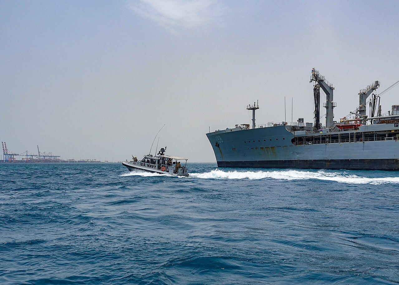 A US Navy patrol boat beside a grey supply ship, with container cranes on the horizon