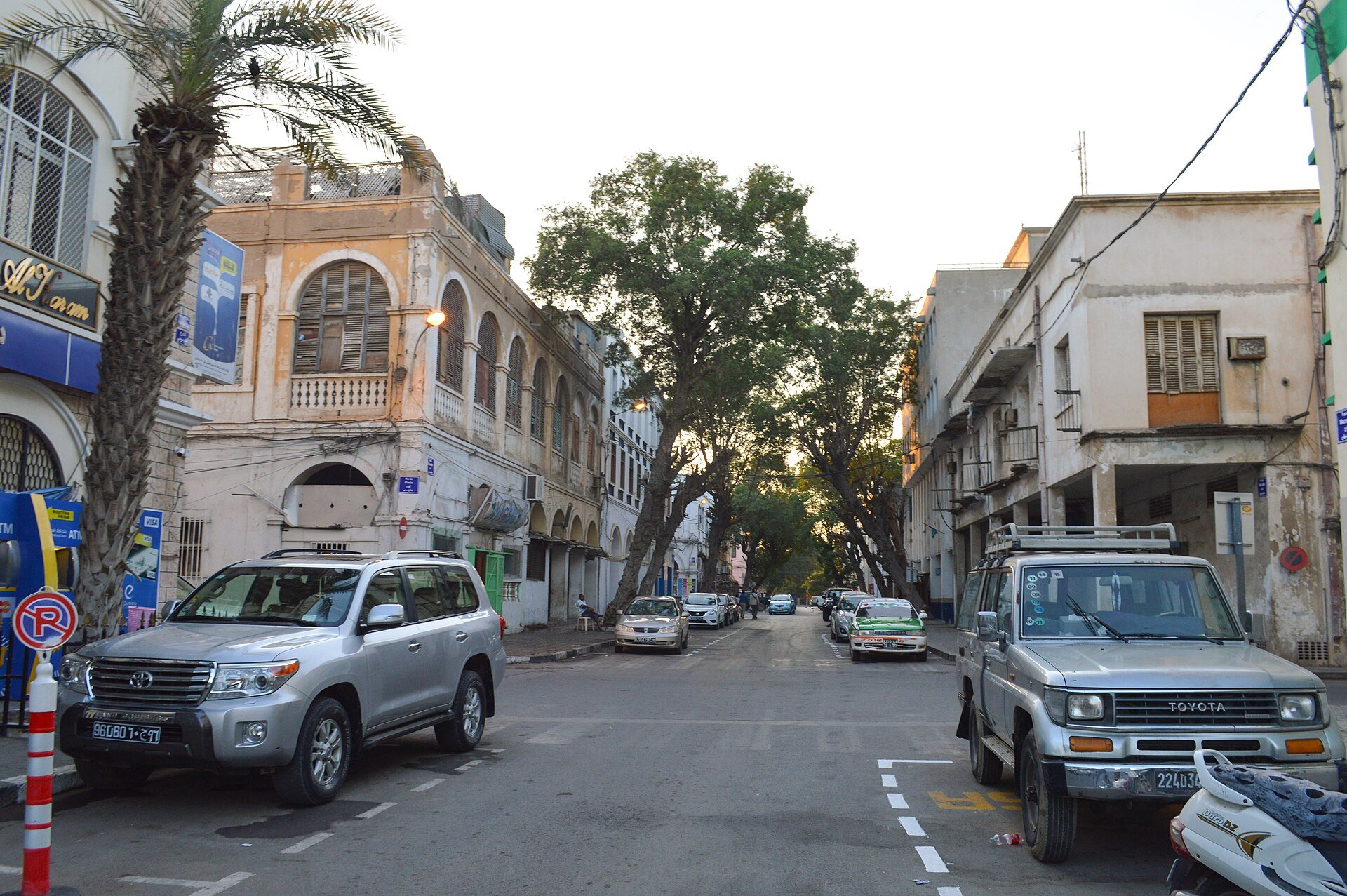 A tree-lined street of older commercial buildings and parked cars in central Djibouti City