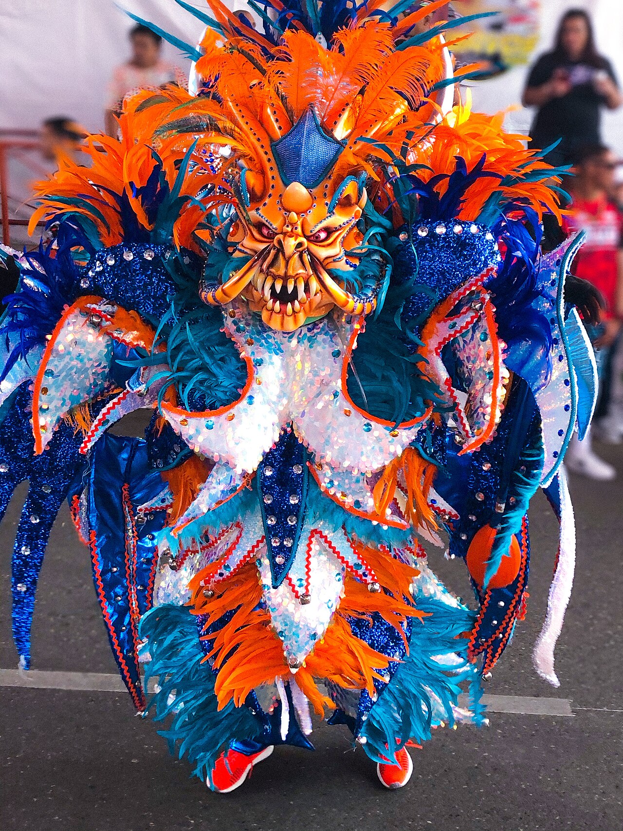 A diablo cojuelo carnival character in an orange and blue feathered costume and fanged mask during the La Vega carnival in the Dominican Republic.