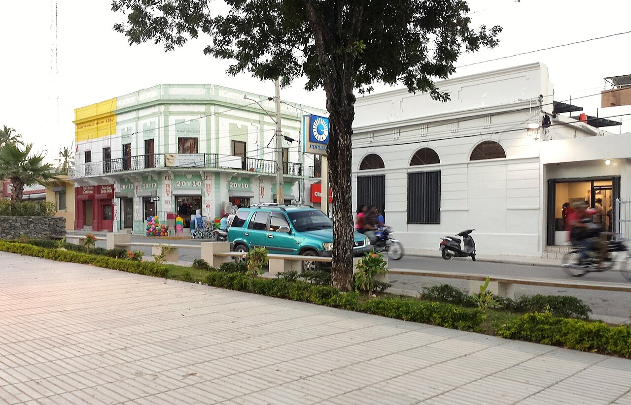 Street corner in downtown Barahona with a pale green two-storey colonial building, a white bank branch, a parked teal SUV and passing motorbikes