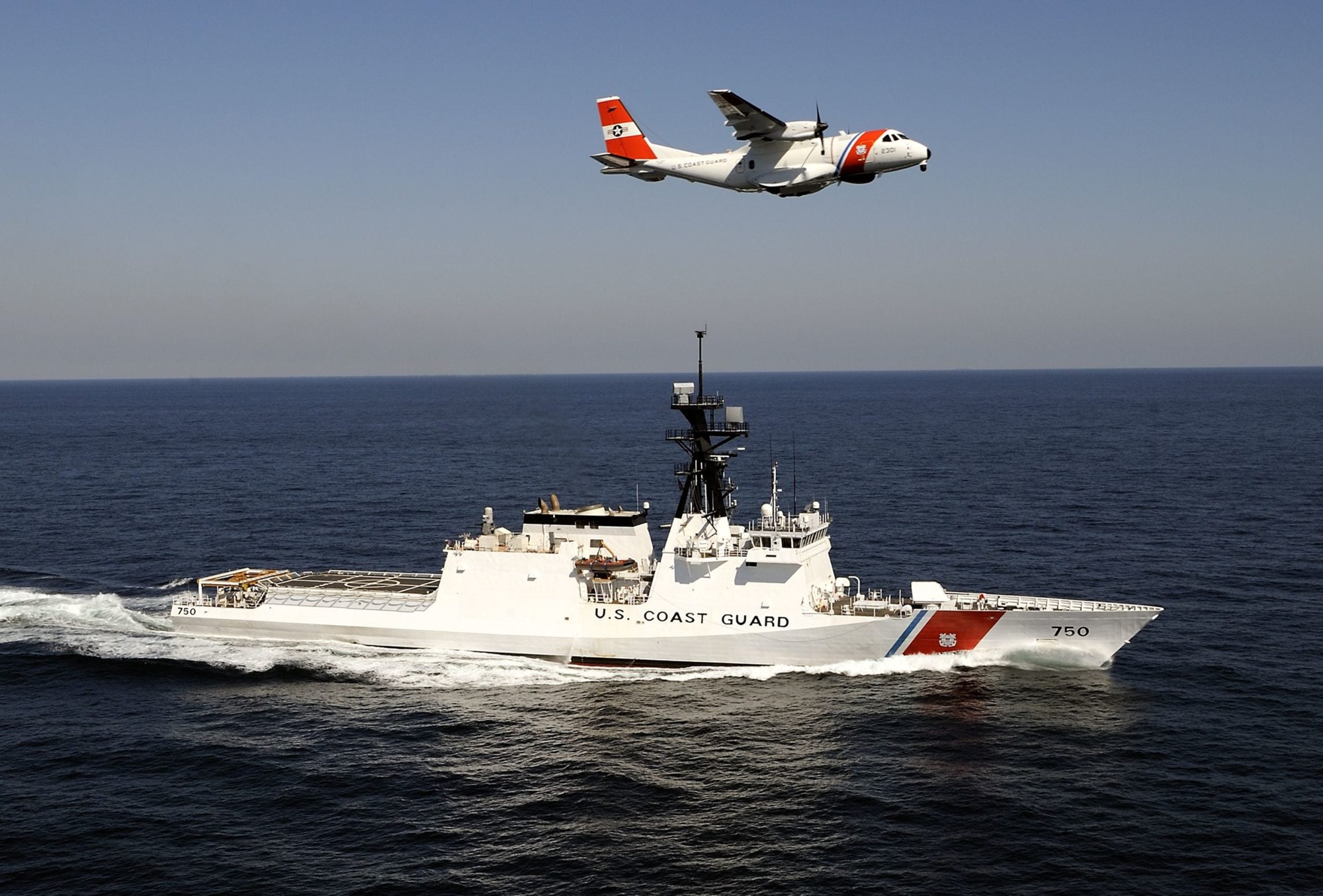 White US Coast Guard cutter at sea with a Coast Guard patrol aircraft flying overhead