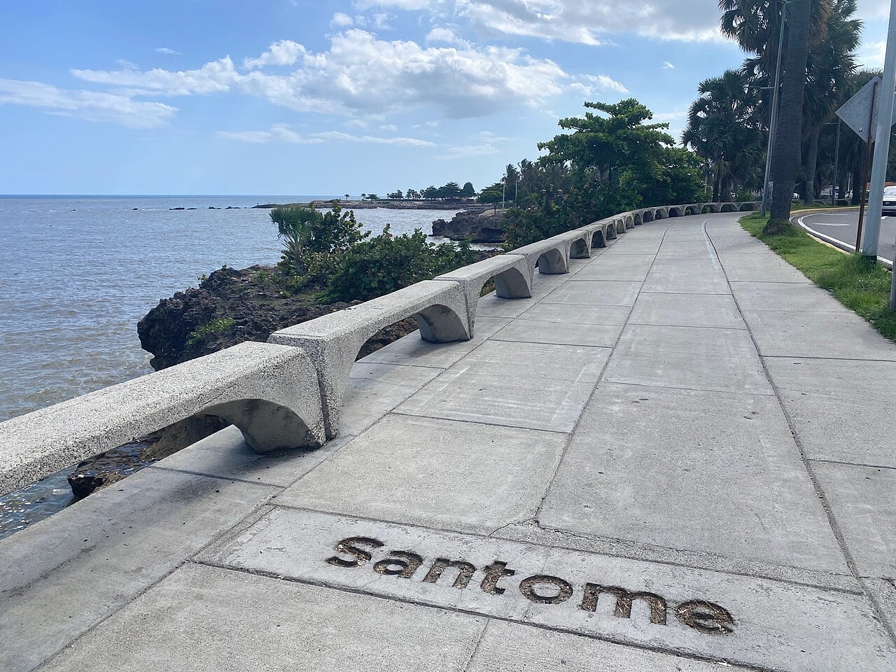 Concrete seafront promenade with curved benches along the rocky Caribbean shore in Santo Domingo, with palm trees and a road beside it