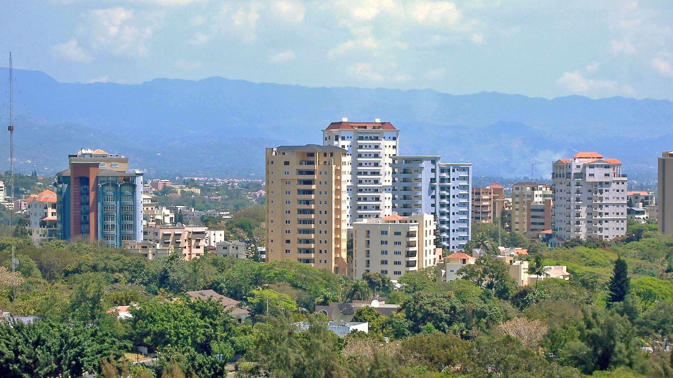 Apartment towers and green treetops in Santiago de los Caballeros with blue mountains behind under a cloudy sky