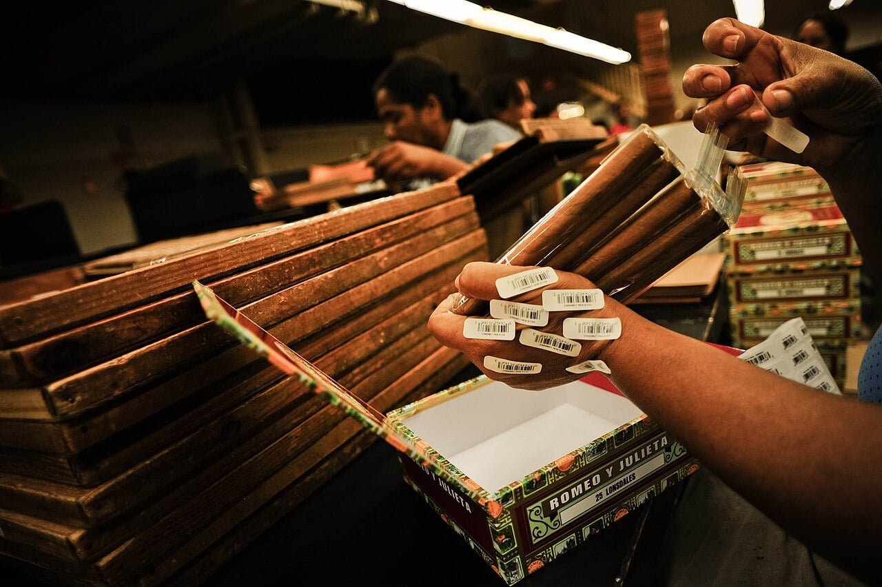 A factory worker holds a bundle of wrapped cigars over an open cigar box, with stacks of boxes behind