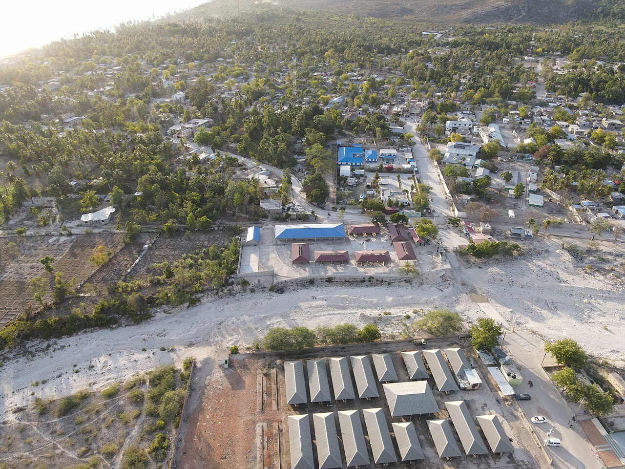 Aerial view of the Haitian town of Anse-à-Pitres beyond a wide dry riverbed, with the grey-roofed market halls of Pedernales, Dominican Republic, in the foreground