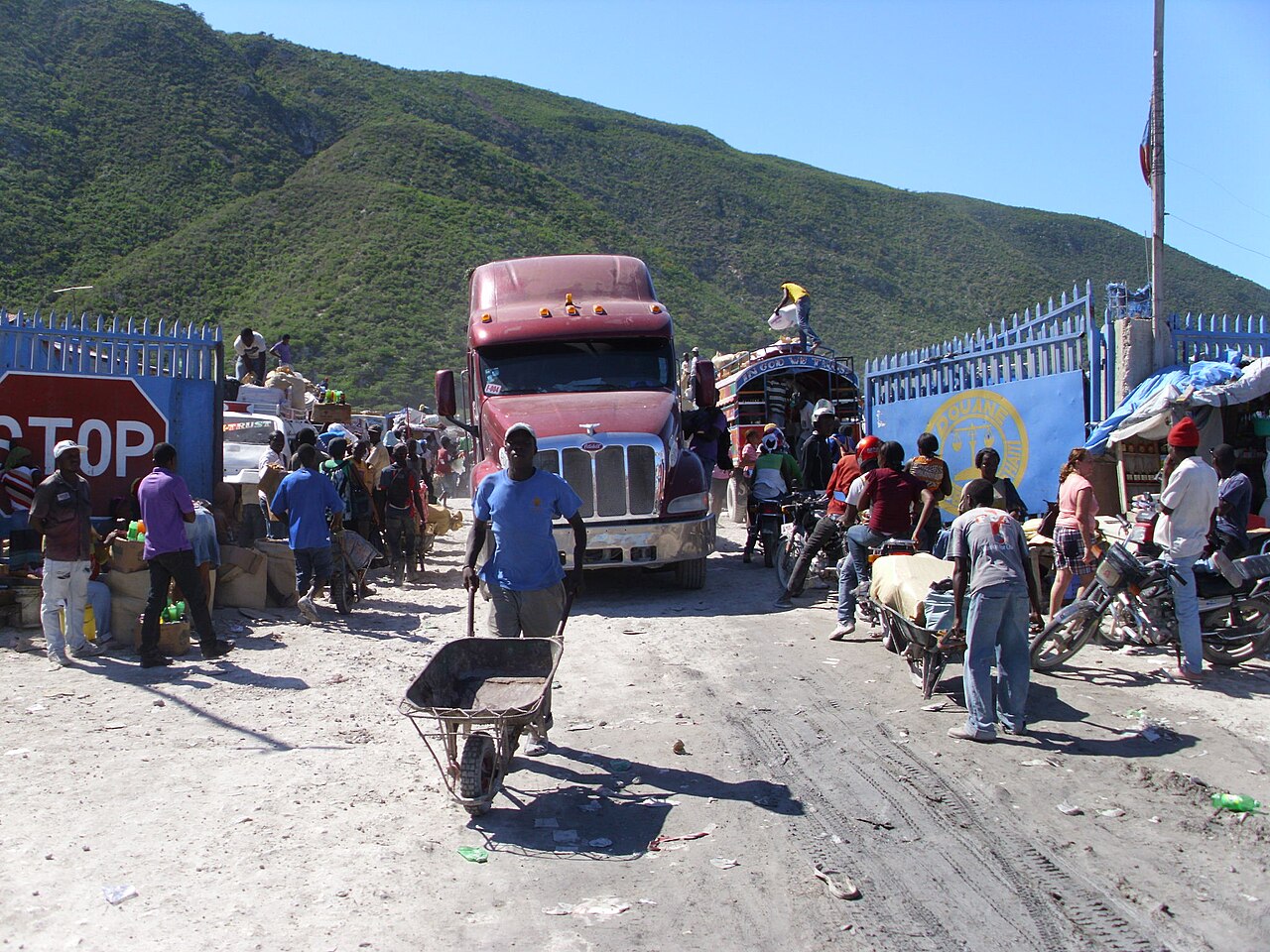 A red lorry, people pushing wheelbarrows and motorcycles crowd through blue customs gates at a dusty border crossing, with green hills behind
