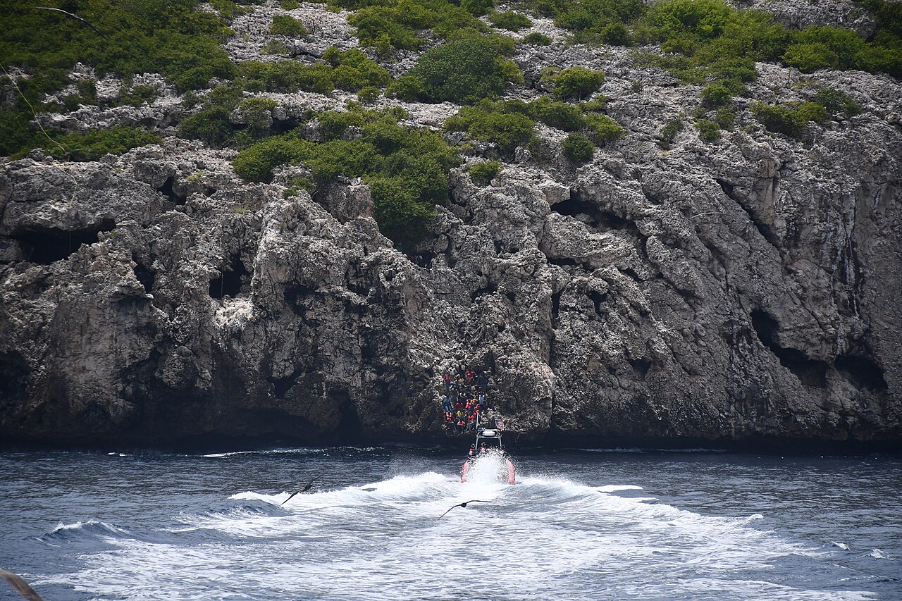 A US Coast Guard rescue boat speeds towards a group of people in orange life jackets clinging to the foot of steep limestone cliffs above the sea