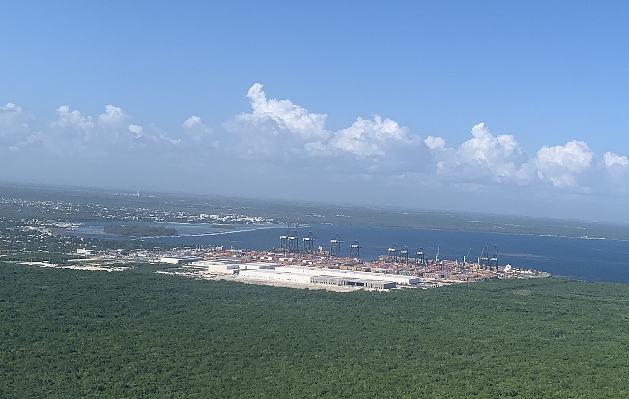 Aerial view of a container port with stacked containers and gantry cranes on a blue bay, surrounded by green forest, with a coastal town behind
