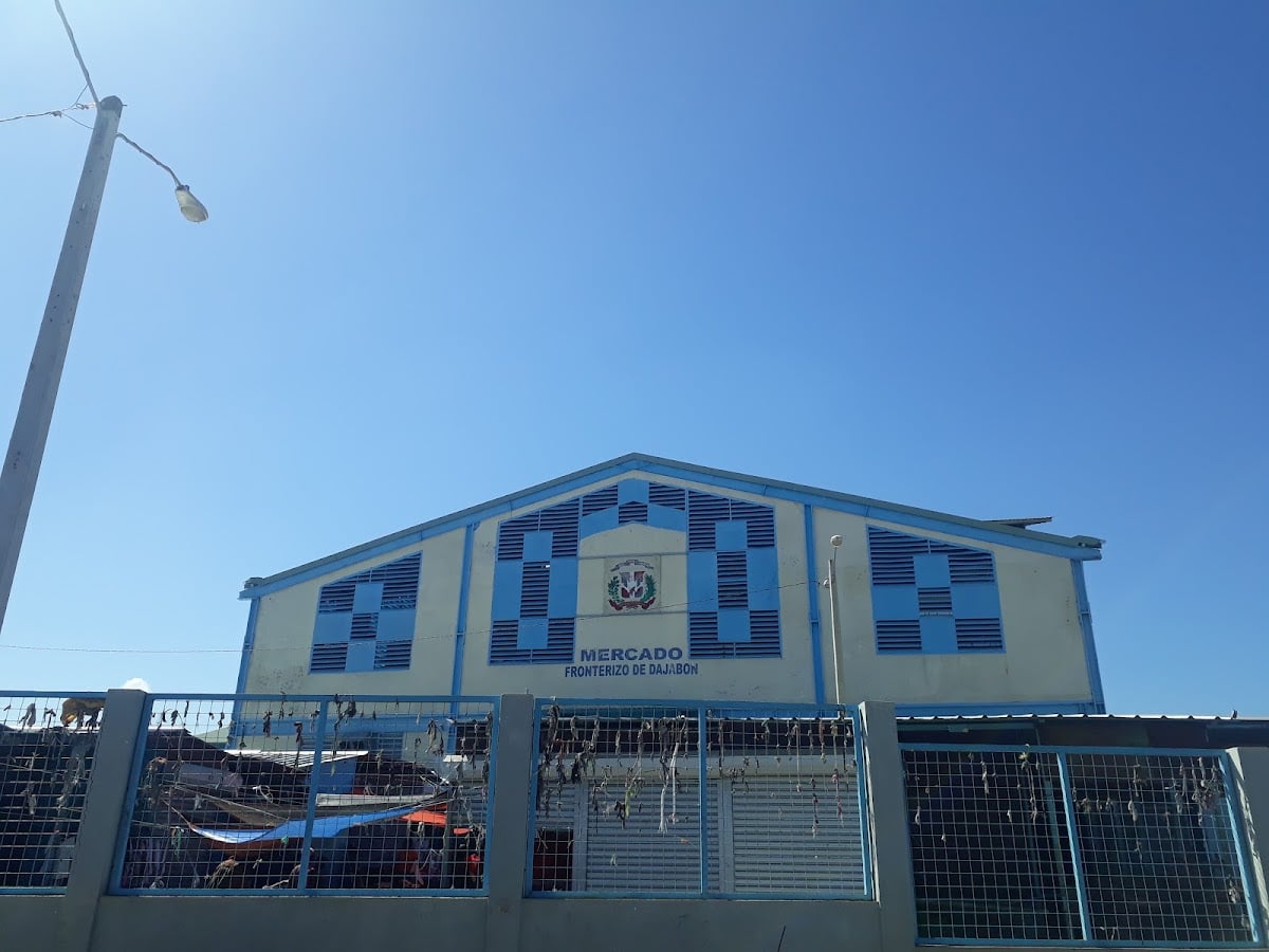 A pale yellow and blue market hall with the Dominican coat of arms and the sign Mercado Fronterizo de Dajabón behind a fence