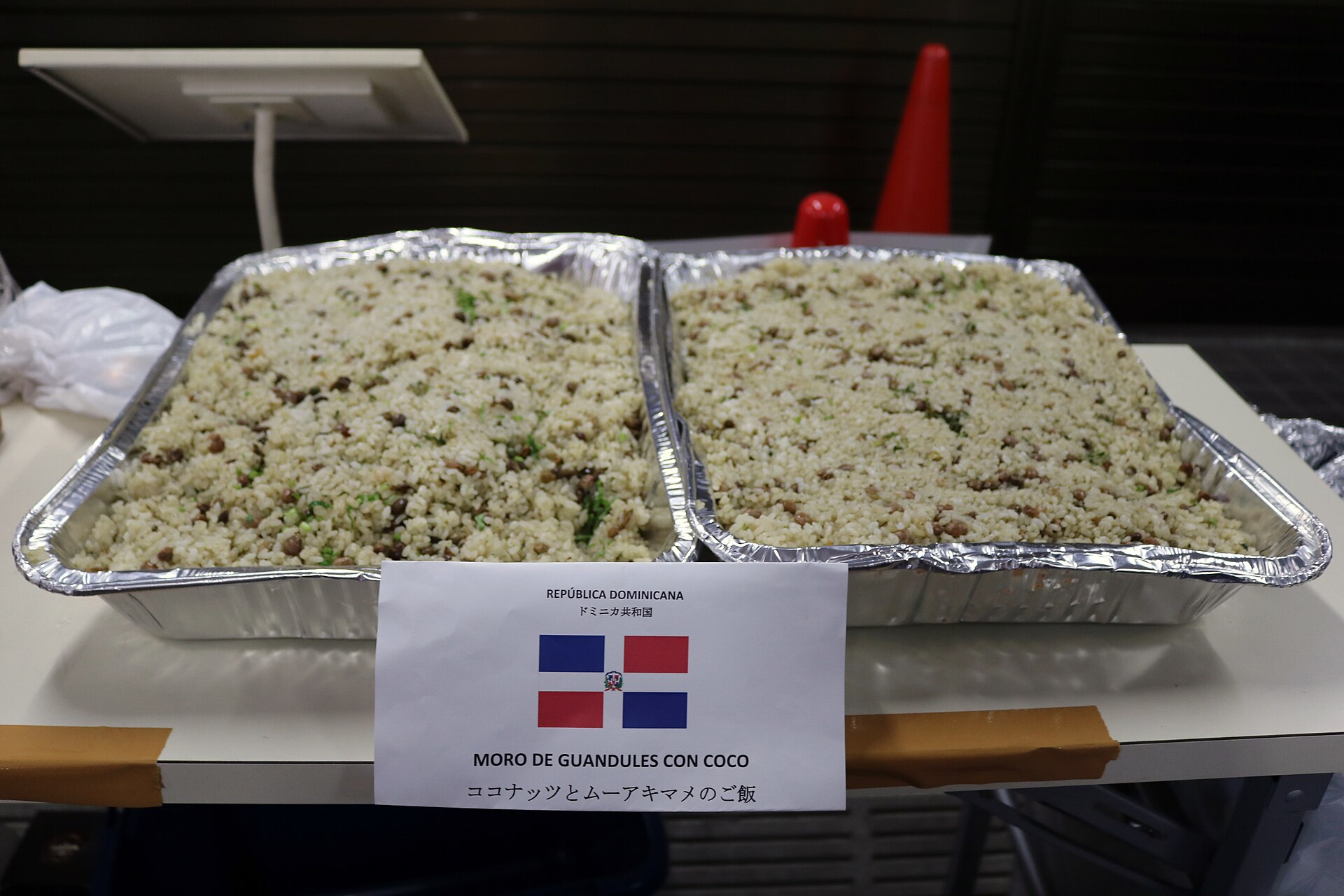 Two foil trays of moro de guandules con coco, a Dominican rice dish, behind a sign with the Dominican flag