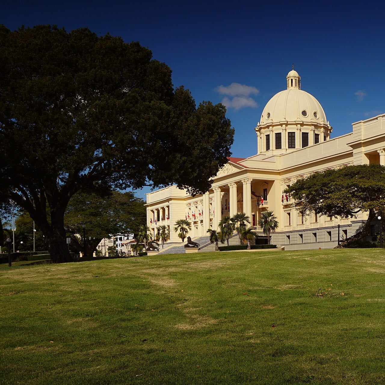 The cream-coloured National Palace in Santo Domingo with its white dome, seen through iron railings across a lawn under a cloudy sky