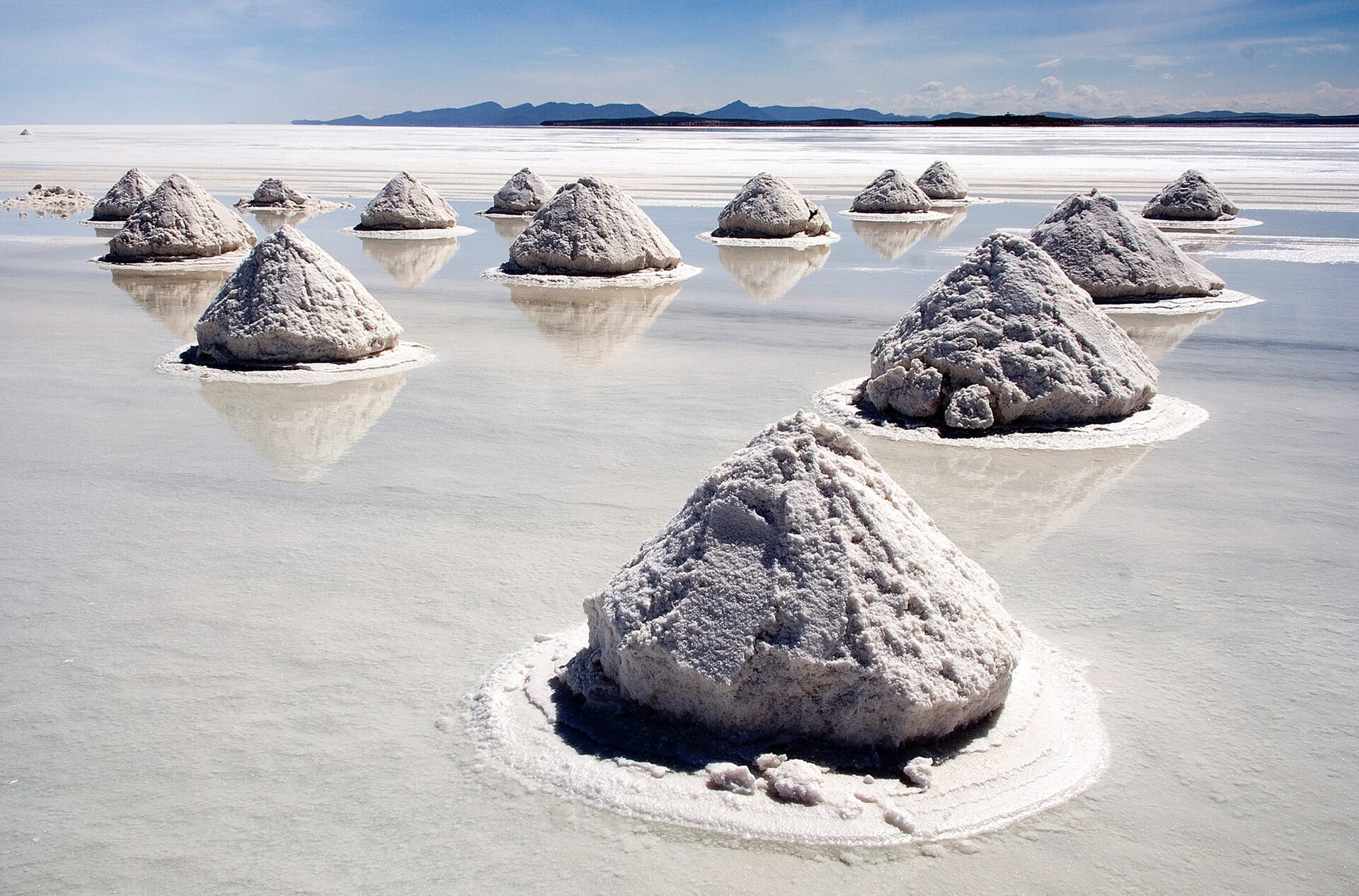 Rows of conical salt piles stand in shallow water on the white Salar de Uyuni, with low mountains on the horizon