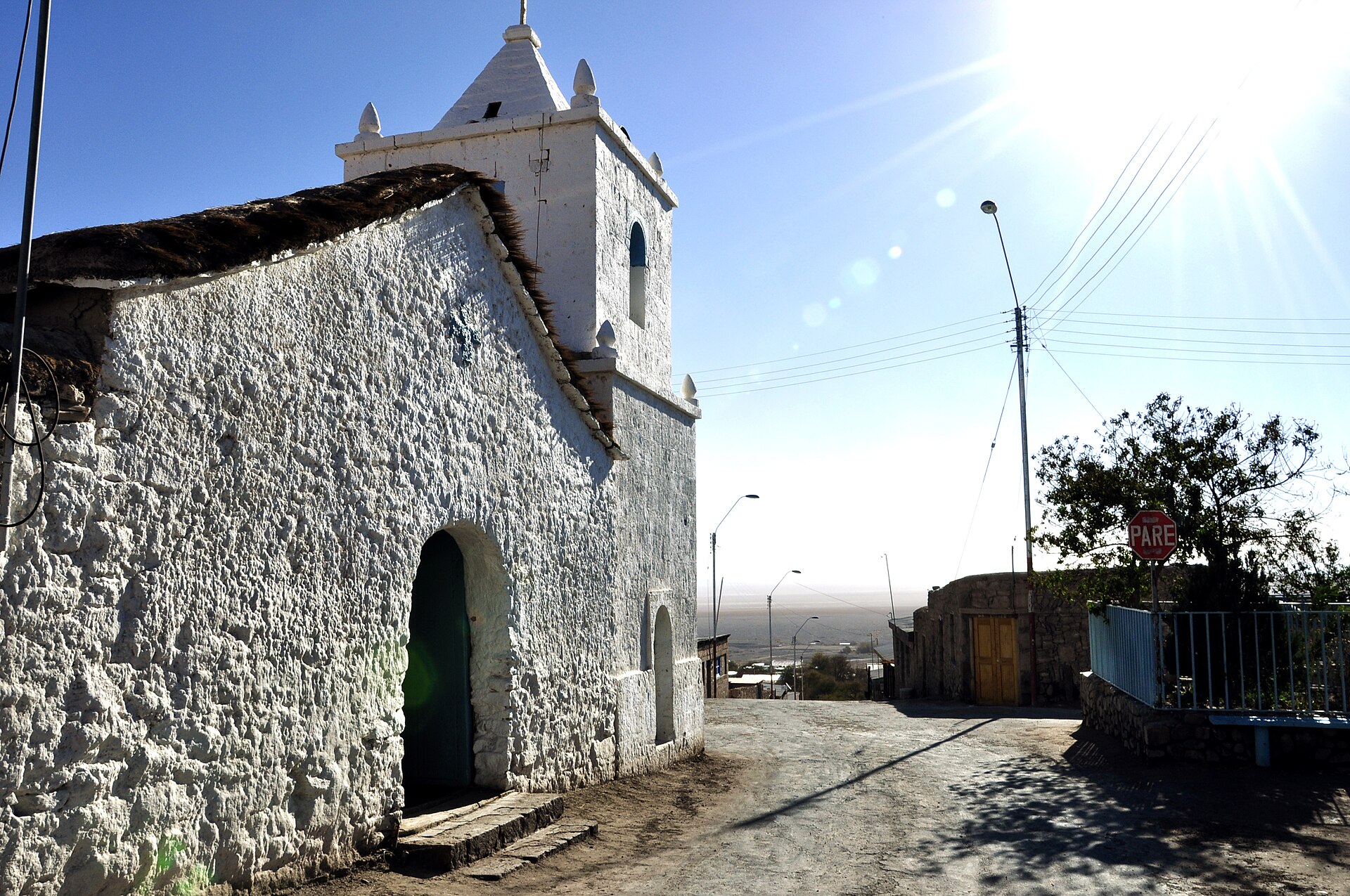 A whitewashed adobe church with a bell tower on a dusty street in the village of Peine, with the flat salt basin visible in the distance