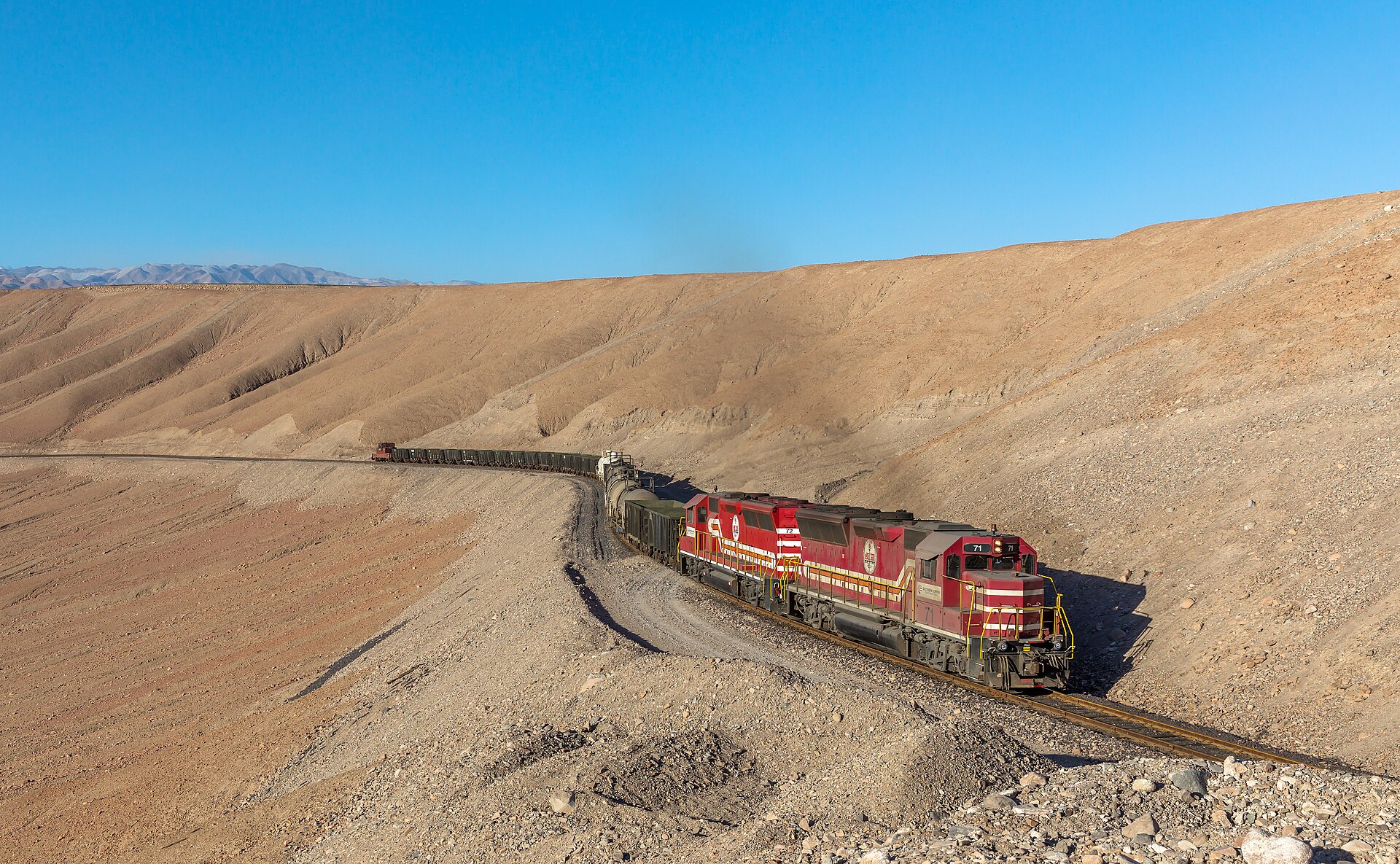 Two red Southern Peru Copper locomotives pull a long freight train around a curve through bare desert hills under a blue sky