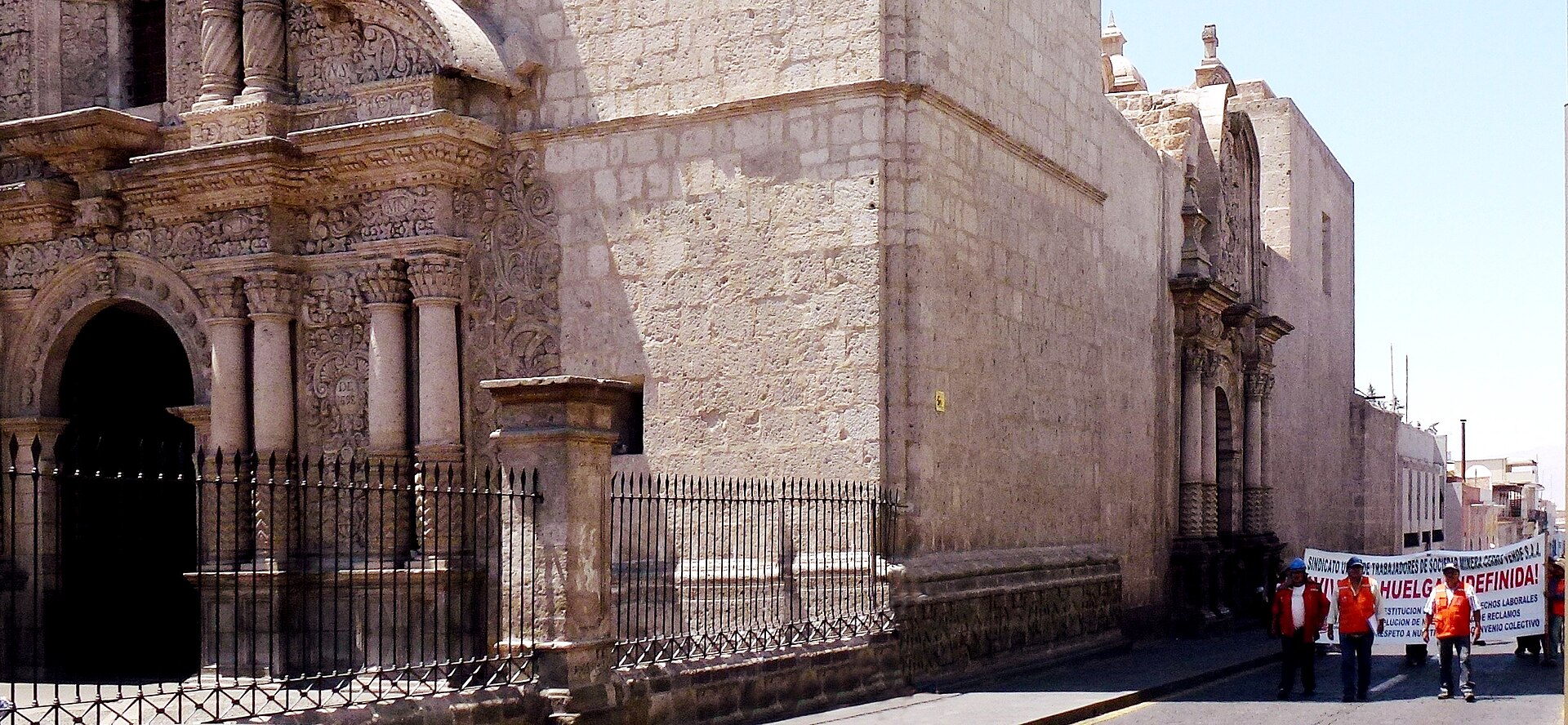 Workers in orange vests carry a banner reading Huelga indefinida past the carved stone facade of a colonial church in Arequipa