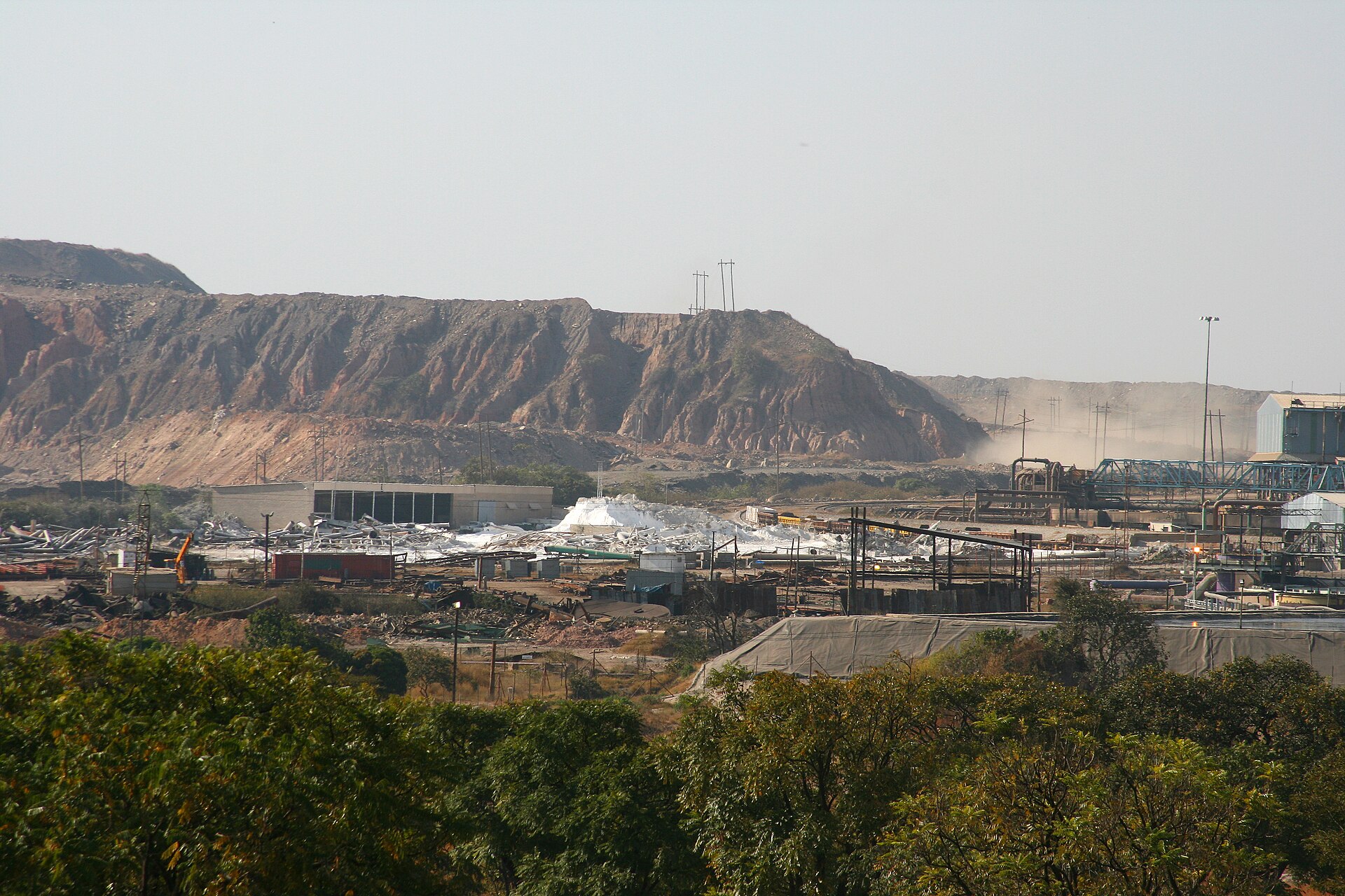 Industrial buildings, conveyors and a white stockpile at a copper mine, with a large brown waste dump behind and trees in the foreground