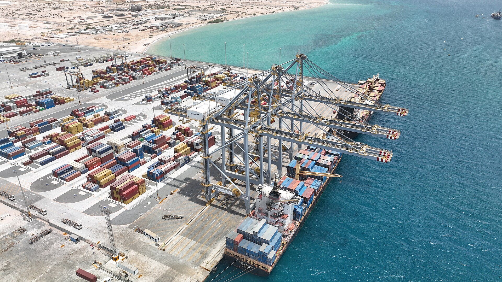 Aerial view of the Berbera container terminal with stacked containers, three ship-to-shore cranes and a loaded container ship beside turquoise water