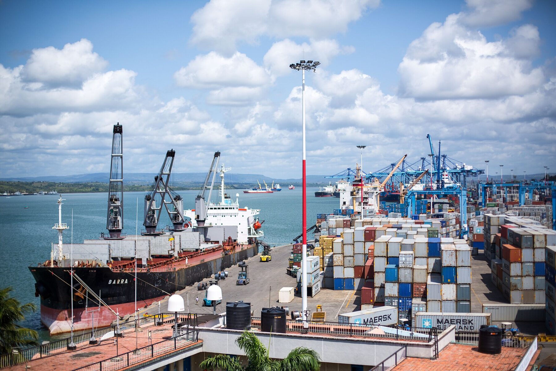 A bulk carrier moored at a quay with cranes, stacks of shipping containers and blue gantry cranes at Mombasa port under a partly cloudy sky, with the harbour behind
