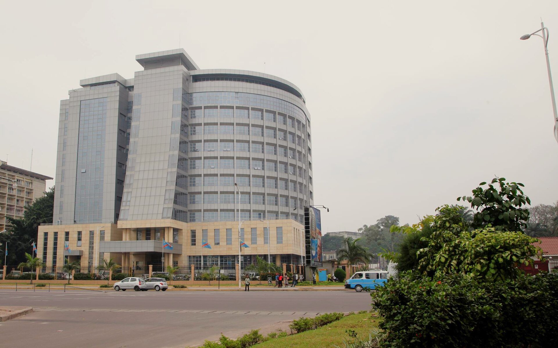 A government office tower in Kinshasa with Congolese flags flying in front of the entrance and cars on the road outside