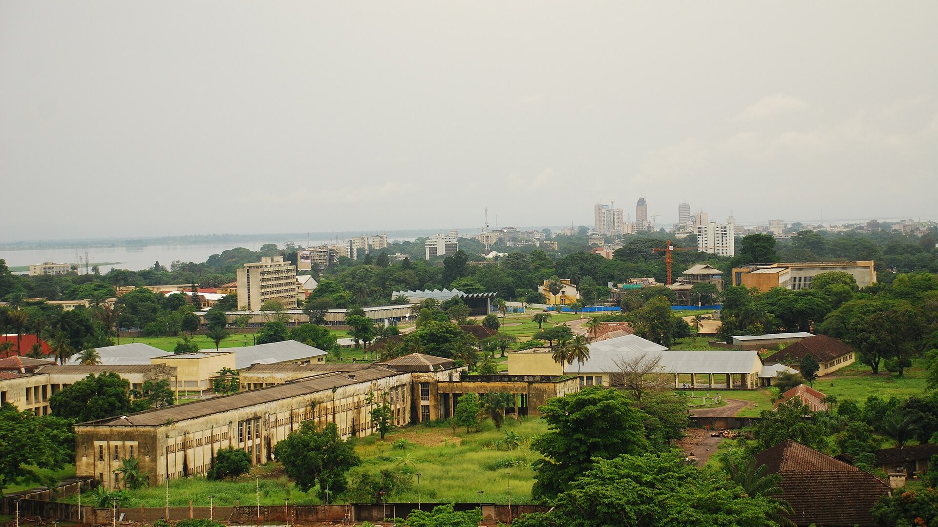 View across Kinshasa's green neighbourhoods and older buildings toward the city centre towers and the Congo River in the haze