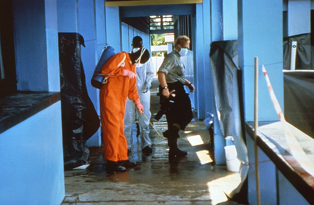 Health workers in orange protective suits and masks disinfect a blue-walled clinic corridor with a sprayer