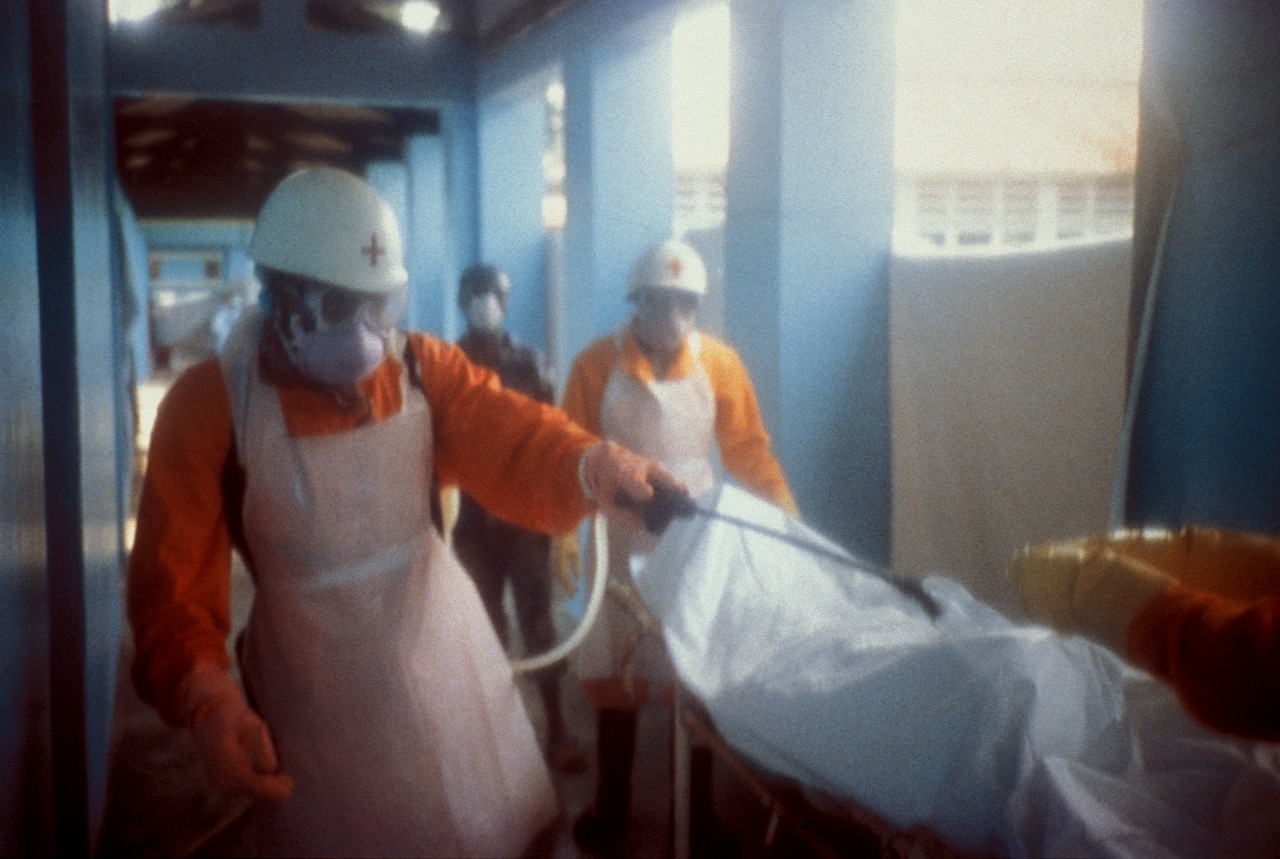 Two Red Cross workers in white helmets, masks and orange sleeves spray a covered stretcher in a clinic corridor