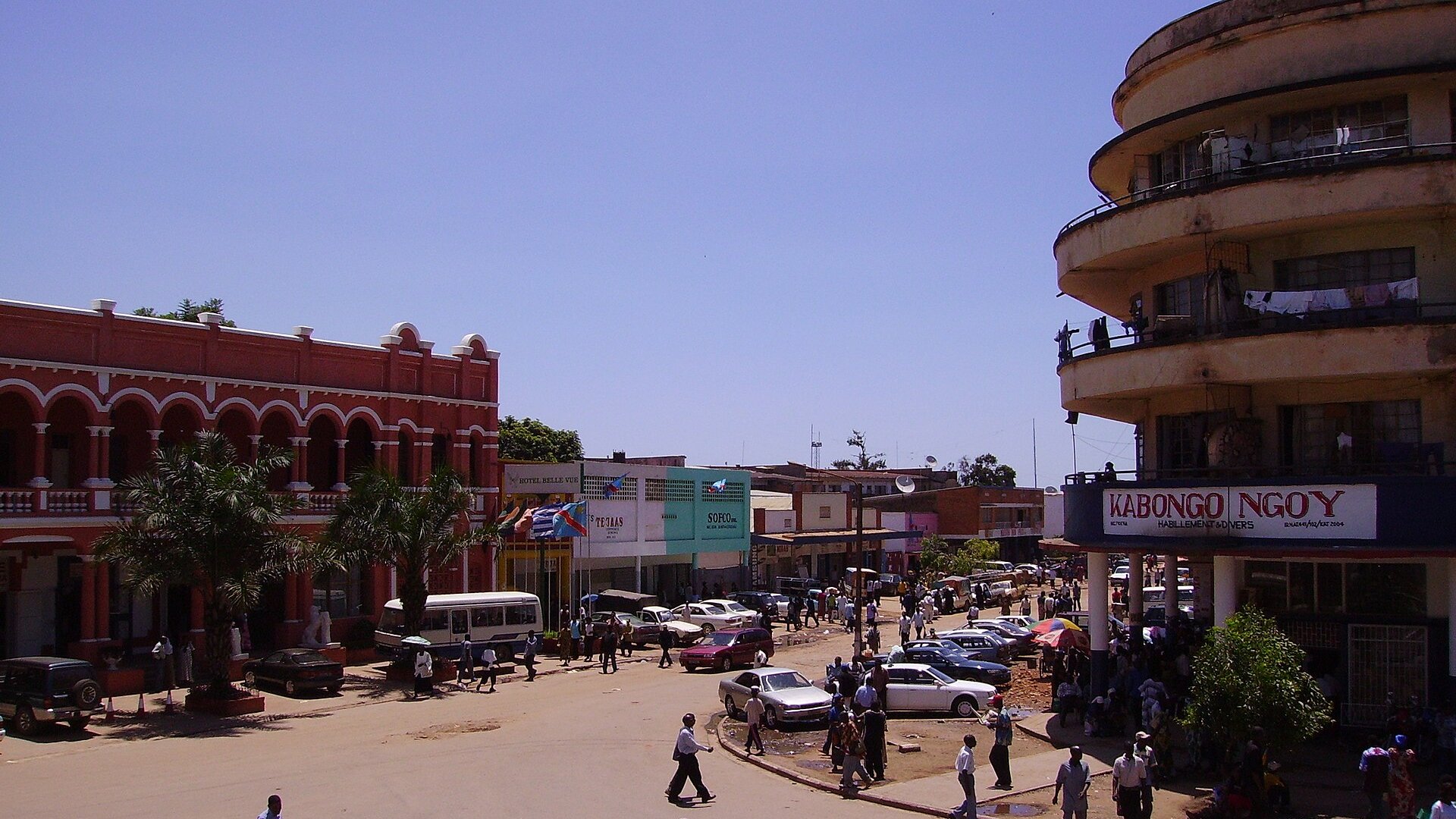 A busy street in downtown Lubumbashi with a red colonial building, shops, cars and pedestrians under a blue sky, DR Congo